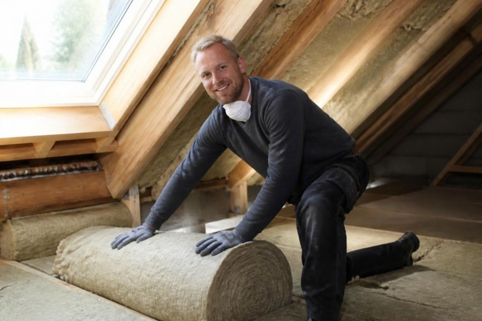 Smilende håndværker med støvmaske om halsen ruller en måtte af mineraluld ud på et loft under et ovenlysvindue