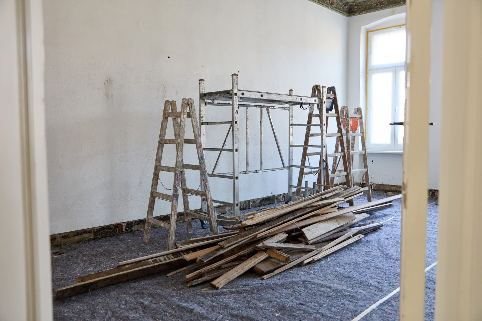 Rullestillads, trappestiger og brædder i et lyst rum med afdækket gulv under renovering
