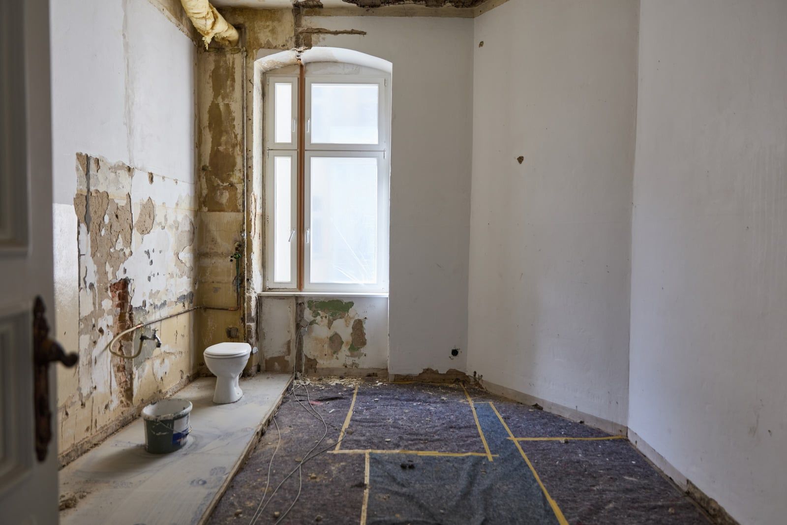 Rum i en ældre lejlighed under renovering med højt vindue, afdækket gulv og afrenset puds